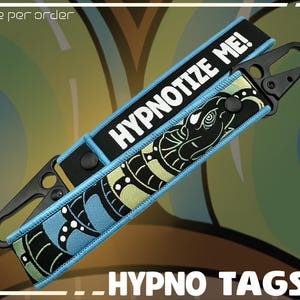 May include: Two black and blue Hypno Tags with a black clasp. One tag reads "HYPNOTIZE ME!" in white text. The other tag features a black and green snake design. The words "_HYPNO TAGS" are at the bottom of the image.