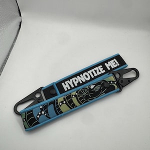 May include: Two keychains with black carabiner clips. One keychain has the text "HYPNOTIZE ME!" in white on a black background. The other keychain has a snake design in black, blue, and green.
