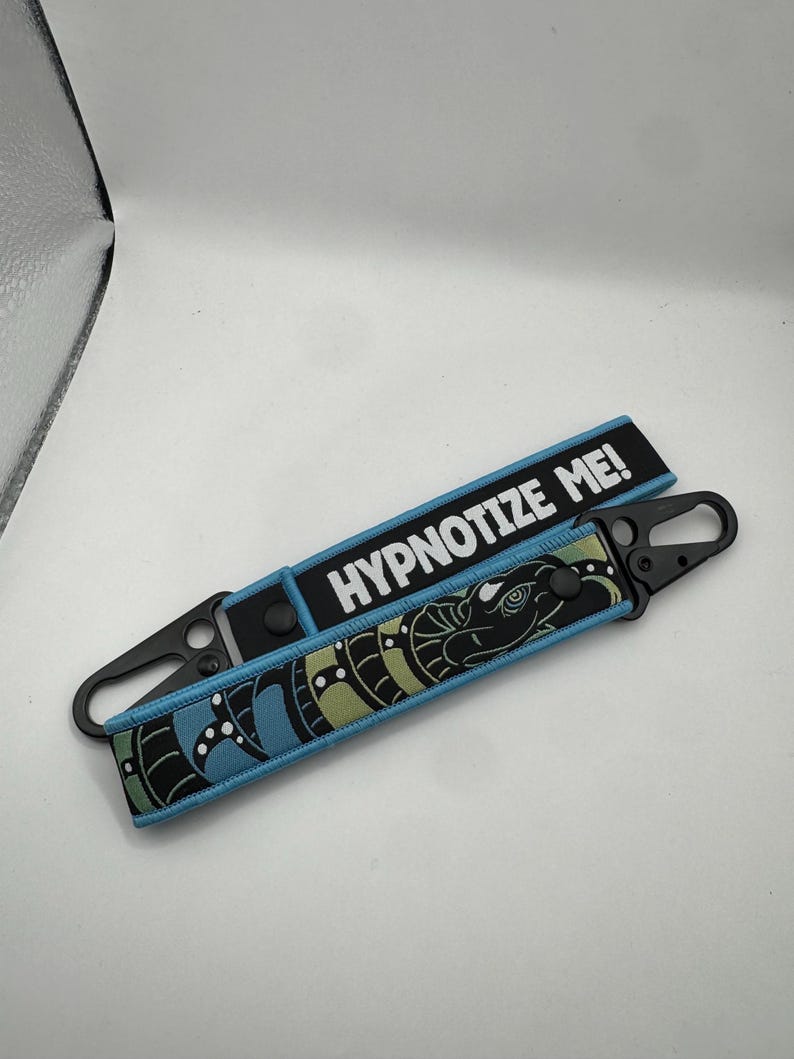 May include: Two keychains with black carabiner clips. One keychain has the text "HYPNOTIZE ME!" in white on a black background. The other keychain has a snake design in black, blue, and green.