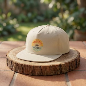 May include: A beige baseball cap with a flat peak, displaying a circular graphic with the text "SEE YOU UP NORTH". The graphic features a sun and wave design in yellow, orange, and blue. The cap is presented on a wooden slice.