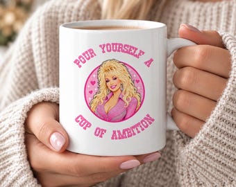 May include: A white ceramic mug featuring a circular pink graphic of a blonde woman with voluminous hair wearing a pink shirt, surrounded by small pink hearts. The text "POUR YOURSELF A CUP OF AMBITION" is printed in pink around the circular graphic.