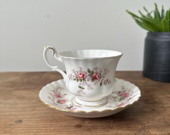 Puede incluir: Una taza de té y un platillo blancos con un patrón floral con rosas rosadas y hojas verdes. El platillo tiene un borde dorado.
