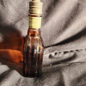 Pu&ograve; includere: Una piccola bottiglia di vetro marrone con un design a coste. La bottiglia ha un tappo di metallo e un'etichetta che recita "1.5 fl oz". La bottiglia &egrave; probabilmente per profumo o un liquido simile.