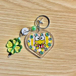 以下が含まれることがあります： ハート型の透明アクリルキーホルダーで、中に黄色いカエルのキャラクターが入っています。カエルは赤と白のストライプのシャツを着ています。キーホルダーには緑の四つ葉のクローバーのチャームとシルバーのキーリングが付いています。