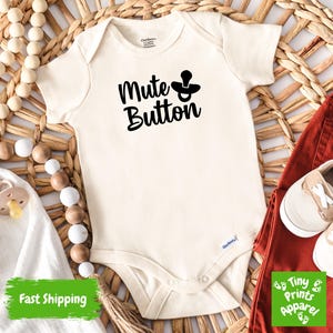 May include: A white baby onesie with black text that reads "Mute Button" and a black pacifier graphic. The onesie is on a woven straw background with a wooden bead garland.