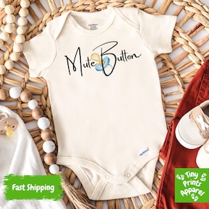 May include: Cream-colored baby onesie with the text "Mute Button" and a pacifier graphic. The onesie is paired with rust-colored pants and small beige and white shoes. The items are arranged on a woven basket.