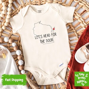 May include: Cream-colored baby onesie with the text "LET'S HEAD FOR THE DOOR" and a Wisconsin outline. The onesie is paired with rust-colored pants and beige baby shoes. A pacifier and wooden beads are also in the image.