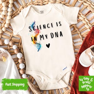 May include: Cream-colored baby onesie with the text "SCIENCE IS IN MY DNA" and a DNA strand graphic. The onesie is paired with rust-colored pants and beige baby shoes. A pacifier and wooden beads are also in the image.