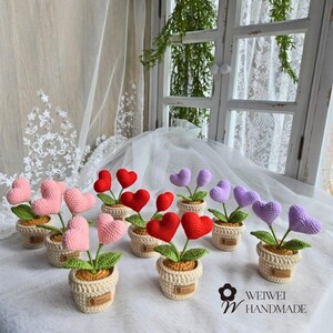 May include: A collection of crocheted heart-shaped flower arrangements in pink, red, and purple. Each arrangement is potted in a small, cream-colored pot and features two hearts on each stem. The pots are arranged on a white surface with a white sheer fabric in the background.
