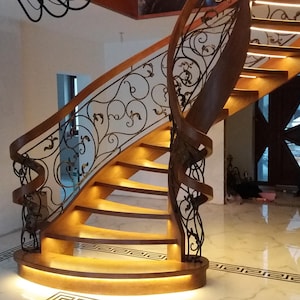 Może przedstawiać: Drewniana klatka schodowa z balustradą z kutego żelaza z kwiatowym wzorem. Schody mają zakrzywiony kształt i są oświetlone ciepłymi białymi światłami.
