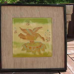 Puede incluir: Un azulejo enmarcado con un fondo verde, amarillo y marrón que presenta un pájaro estilizado con un cuerpo marrón y alas verdes. El pájaro está posado sobre un diseño floral verde y amarillo.