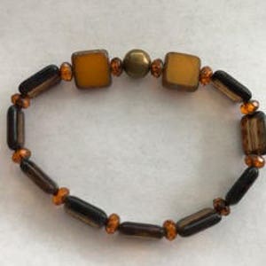 May include: A brown and amber beaded bracelet with a gold-toned bead in the center. The bracelet features square-shaped beads.