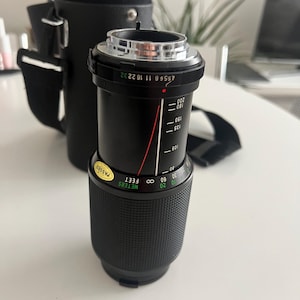 Peut inclure: Un objectif d'appareil photo noir avec une bague argentée et un point rouge. L'objectif a une échelle de profondeur de champ marquée en mètres et en pieds. L'objectif est marqué "PASSED".