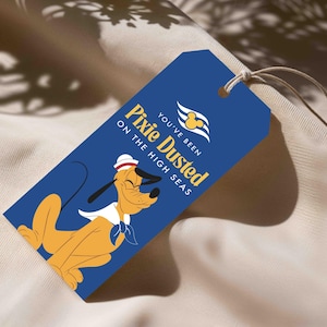 Op de afbeelding: Een rechthoekig blauw label met een touwtje, voorzien van een gele cartoonhond met een witte matrozenmuts en een blauwe halsdoek. De tekst luidt YOU'VE BEEN PIXIE DUSTED ON THE HIGH SEAS met een geel Mickey Mouse-icoon erboven.
