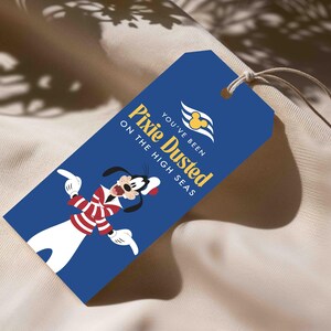 Op de afbeelding: Een rechthoekig blauw cadeaulabel met een touwtje, voorzien van een afbeelding van een stripfiguurhond in een rood-wit gestreept shirt. De tekst luidt: You've been Pixie Dusted on the High Seas, met een geel Mickey Mouse-logo.