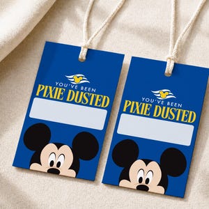Puede incluir: Dos etiquetas de equipaje rectangulares azules con cordones blancos. Cada etiqueta presenta un icono amarillo de la cabeza de Mickey Mouse, el texto "YOU'VE BEEN PIXIE DUSTED" en amarillo, un espacio blanco para escribir y la cara de Mickey Mouse.