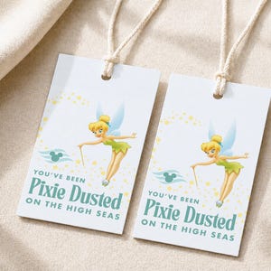 Op de afbeelding: Twee rechthoekige witte papieren labels met crèmekleurige touwtjes. Elk label toont een blonde fee in een groene jurk met blauwe vleugels, gele glitters, een turquoise Mickey Mouse-icoon en de tekst You've Been Pixie Dusted On The High Seas.