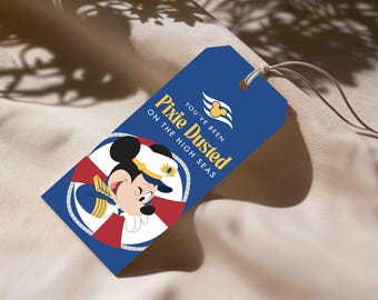 Puede incluir: Una etiqueta de regalo rectangular azul con una cuerda, que presenta un gráfico de Mickey Mouse con uniforme de capitán dentro de un salvavidas rojo y blanco. El texto dice "YOU'VE BEEN PIXIE DUSTED ON THE HIGH SEAS" con un logo de crucero.