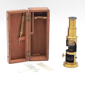 Puede incluir: Un microscopio de latón antiguo con una caja de almacenamiento de madera. El microscopio es vertical con un cuerpo cilíndrico y perillas ajustables. La caja de madera abierta contiene un instrumento de latón y un compartimento. También hay varias diapositivas de vidrio.