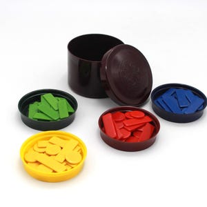 Puede incluir: Un recipiente cilíndrico marrón oscuro con una tapa a juego, rodeado de cuatro pequeños recipientes abiertos, negros y amarillos. Cada recipiente pequeño contiene piezas de juego de diferentes colores: verde, rojo, azul y amarillo.