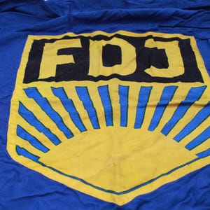 May include: A blue fabric with a yellow and blue emblem. The emblem features the letters "FDJ" in black, set against a yellow sunburst design with blue rays. The fabric is wrinkled.