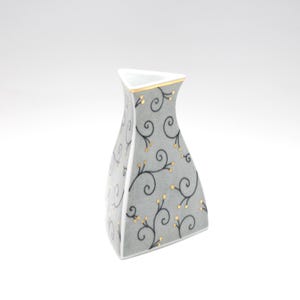 May include: A grey ceramic vase with a distinctive shape and a decorative floral pattern. The vase features black swirling vines with small gold accents. The rim is trimmed with gold. The vase is approximately 15 cm tall.