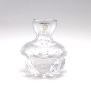 Könnte beinhalten: Eine klare Glasvase mit einem einzigartigen, strukturierten Design. Die Vase hat einen bauchigen Boden mit einer strukturierten, blütenartigen Form und einen schlanken Hals. Ein kleines Etikett ist oben sichtbar. Die Vase dient wahrscheinlich dekorativen Zwecken.