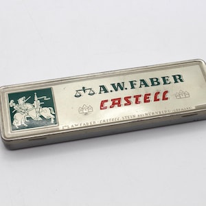 Op de afbeelding: Een rechthoekige metalen etui met een zilveren afwerking. De bovenkant is voorzien van de tekst "A.W. FABER CASTELL" in groen en rood, samen met een groen-witte illustratie van een ridder te paard. De etui is waarschijnlijk vintage.