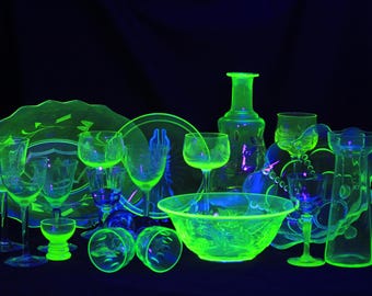 May include: A collection of various glass items including plates, bowls, a decanter, and stemmed glasses, all glowing with a bright neon green hue under ultraviolet light, arranged against a dark background to highlight their fluorescent properties.