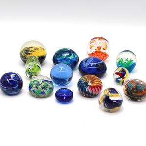 Könnte beinhalten: Eine Sammlung dekorativer Glasmurmeln in verschiedenen Größen und Farben. Die Murmeln weisen wirbelnde Muster und eingebettete Designs in Blau-, Grün-, Gelb-, Orange- und Rottönen auf. Einige sind klar, andere undurchsichtig.