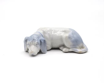 Puede incluir: Figura de cerámica de un perro salchicha en reposo. El perro es principalmente blanco con detalles en azul claro en las orejas y la cabeza. La figura es un objeto decorativo, probablemente para exhibición.