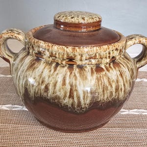 Puede incluir: Un tarro de cerámica esmaltado en marrón y crema con tapa y dos asas. El tarro tiene un esmalte marrón y crema moteado.