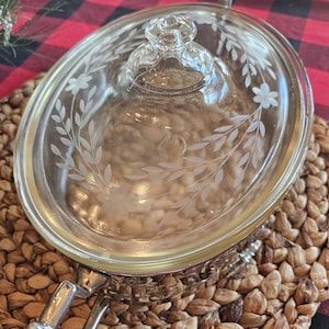 Könnte beinhalten: Eine ovale Auflaufform aus klarem Glas mit einem floralen Ätzdesign auf dem Deckel. Die Form steht in einem silberfarbenen Metallhalter mit Griffen. Der Deckel hat einen Knaufgriff. Die Form steht auf einer gewebten Matte.
