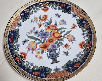 May include: A white ceramic plate with a blue and gold rim. The plate features a floral design with orange, red, and yellow flowers in a vase. The flowers are surrounded by green leaves and butterflies.