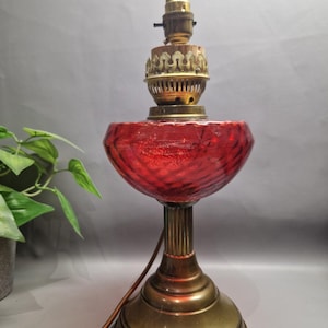 May include: A table lamp featuring a vibrant red glass bowl with a textured, swirling pattern. The lamp has a brass base and a brass burner assembly at the top. A brown electrical cord extends from the base of the lamp, which stands upright.