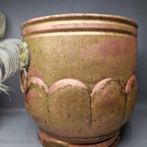 May include: A round, aged copper planter with a weathered patina. The planter features a decorative scalloped design around the middle and a floral accent on the side. The color is a mix of green and pink.