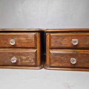 May include: Two small, wooden chests of drawers, each with two drawers. The chests are stained a medium brown and feature crystal-like knobs. The wood grain is visible, suggesting a vintage or antique style. The chests are placed side by side.