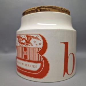 May include: A white ceramic jar with a cork lid. The jar features a large red "B" and a red graphic with an eagle and the words "Beans & Berries". The jar is cylindrical and has a slightly flared top.