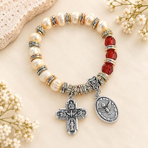 May include: A stretch bracelet featuring cream-colored freshwater pearls, three faceted translucent red beads, and silver-toned metal spacers with clear crystals. Two silver-toned metal charms hang from the bracelet: a cross and an oval medal.
