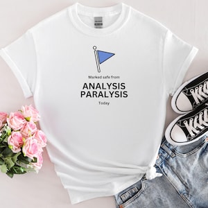 May include: White t-shirt with the text "Marked safe from ANALYSIS PARALYSIS Today" and a blue flag graphic. The shirt is paired with blue jeans and black and white sneakers. A bouquet of pink roses is in the background.