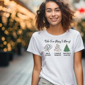 以下が含まれることがあります： 「Data: From Messy to Merry!」というテキストとクリスマスツリーのデザインが施された白いTシャツ。デザインは、データ分析の異なる段階を表す、ライトが付いたクリスマスツリーが3本描かれています。生のままの状態、整理された状態、分析と視覚化された状態です。