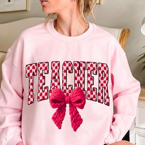 Könnte beinhalten: Hellrosa Sweatshirt mit dem Wort "TEACHER" in fetten Blockbuchstaben. Die Buchstaben sind mit einem rot-weißen Herzmuster gefüllt. Eine große, rote Strickschleife befindet sich unter dem Text.