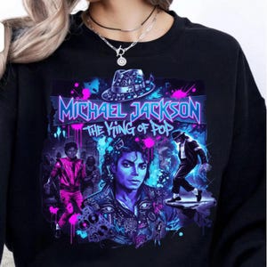 May include: Black sweatshirt featuring a vibrant graphic of Michael Jackson. The design includes the text "Michael Jackson The King of Pop" in blue and pink, along with images of Jackson in various poses. The sweatshirt has a crew neck.