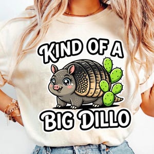 May include: A cream-colored t-shirt featuring a cartoon armadillo with a cactus. The armadillo is gray with a black shell and pink accents. The text "KIND OF A BIG DILLO" is printed above and below the armadillo in black.