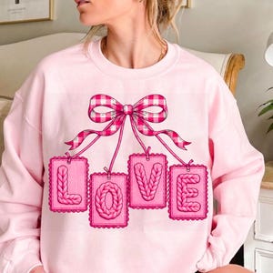 Puede incluir: Sudadera rosa claro con un gran lazo de cuadros rosa y blanco. Debajo del lazo, cuatro etiquetas rectangulares rosas deletrean la palabra "LOVE" en un estilo tejido.