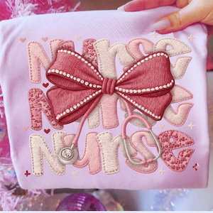 May include: A light pink t-shirt with the word "NURSE" embroidered in a mix of pink and cream colors. A large red bow with white beaded trim and a stethoscope are also embroidered on the shirt. Small hearts and stars are scattered around the design.