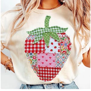 May include: Cream-coloured t-shirt with a large, patchwork strawberry design. The strawberry is made of various fabric patterns, including red gingham, floral prints, and pink hearts. The stem and leaves are green.