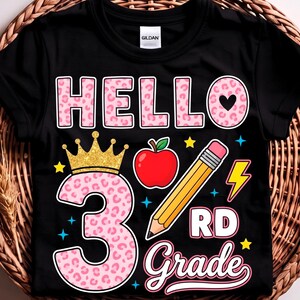May include: A black short-sleeve t-shirt featuring a graphic print that says HELLO 3RD Grade. The design includes a large pink leopard print number 3 topped with a gold crown, a red apple, a yellow pencil, a yellow lightning bolt, and stars.