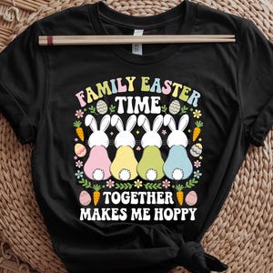 Op de afbeelding: Zwart T-shirt met een afbeelding van vier paashazen die rug aan rug zitten. De tekst "Family Easter Time Together Makes Me Hoppy" staat op het shirt gedrukt.