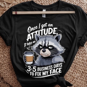 May include: Black t-shirt with a cartoon raccoon holding a coffee cup and a grumpy expression. The text on the shirt reads "Once I get an ATTITUDE, It takes me 3-5 BUSINESS DAYS TO FIX MY FACE". The shirt is made of a soft fabric.